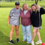 Three women standing on a golf course smiling at the camera.