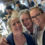 Three women smiling closely together in a lively indoor setting.