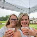 Two women smiling and holding drinks under a white canopy outdoors.