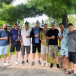 Group of eight men posing outdoors under a tree.