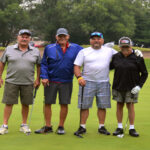 Four men posing on a golf course with greenery in the background.