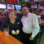 Two people posing at a bar counter with drinks.